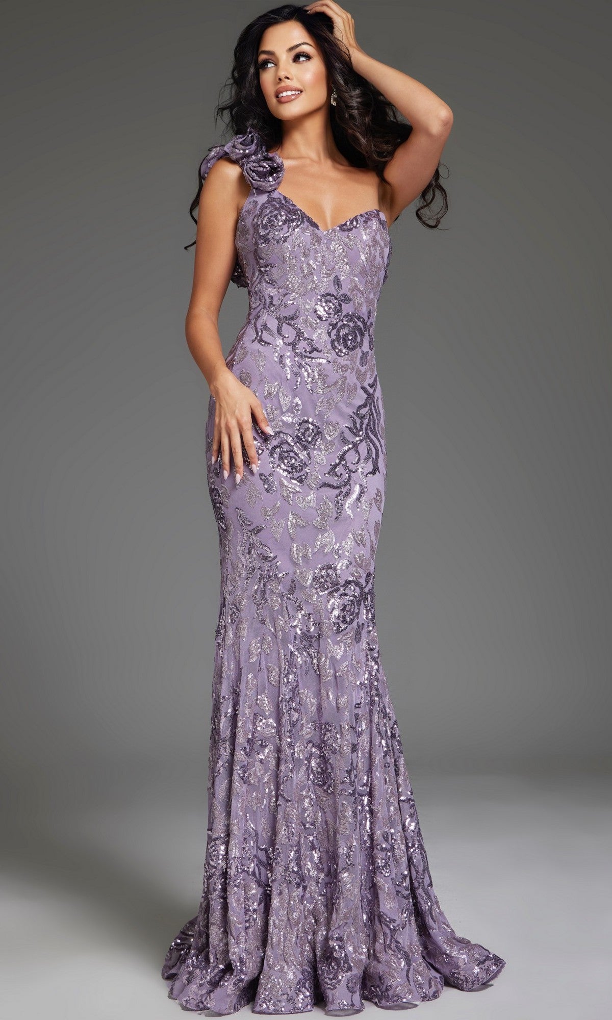Woman wearing Jovani 37634, a fitted, floor-length lavender sequin prom dress with a mermaid silhouette and silver floral accents, posing with one hand on her hip and the other in her hair against a gray background.